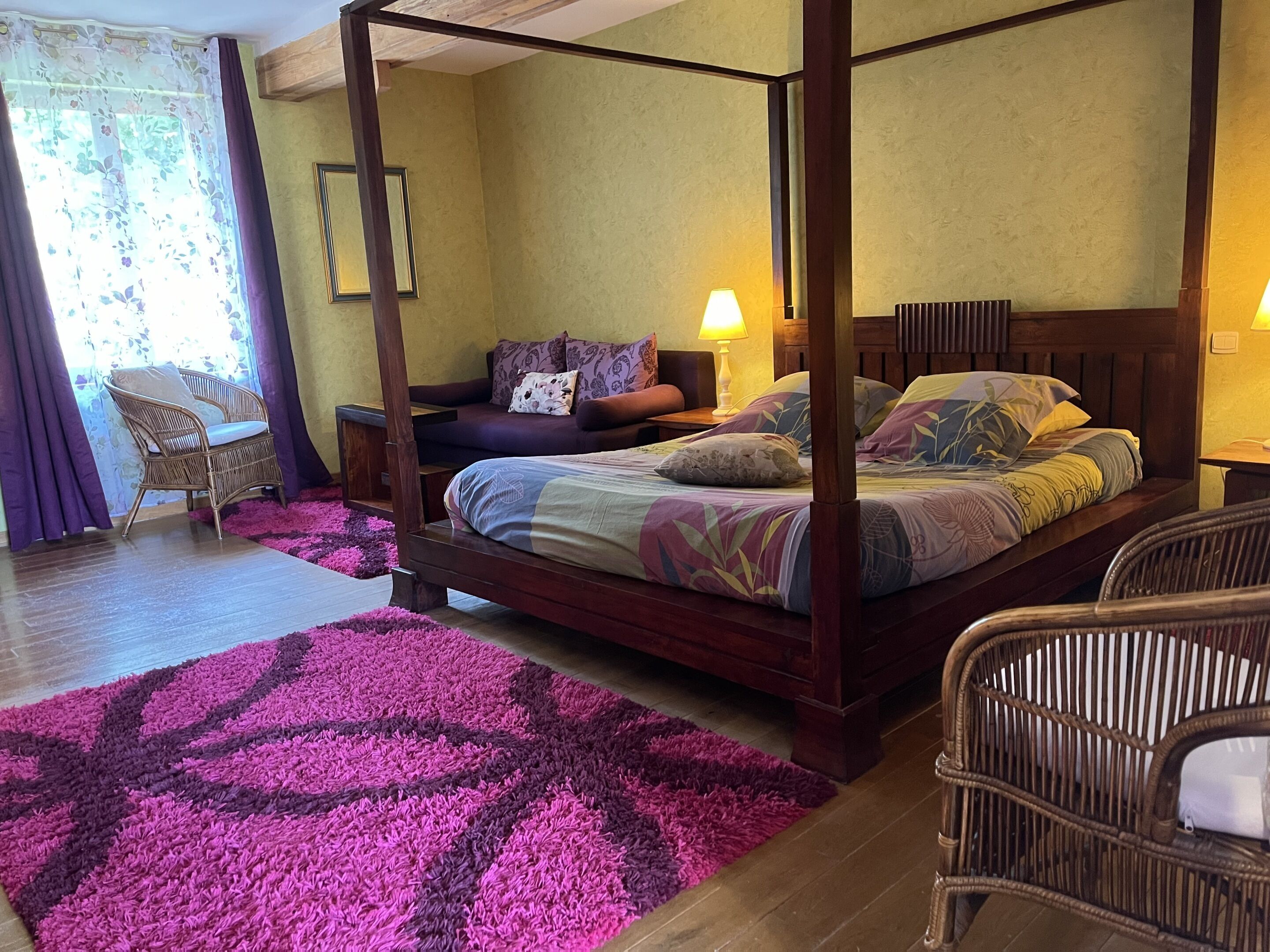 En seng med himmelseng, et blomstret sengetæppe, et lilla og pink tæppe, en fletstol og et natbord med en lampe.