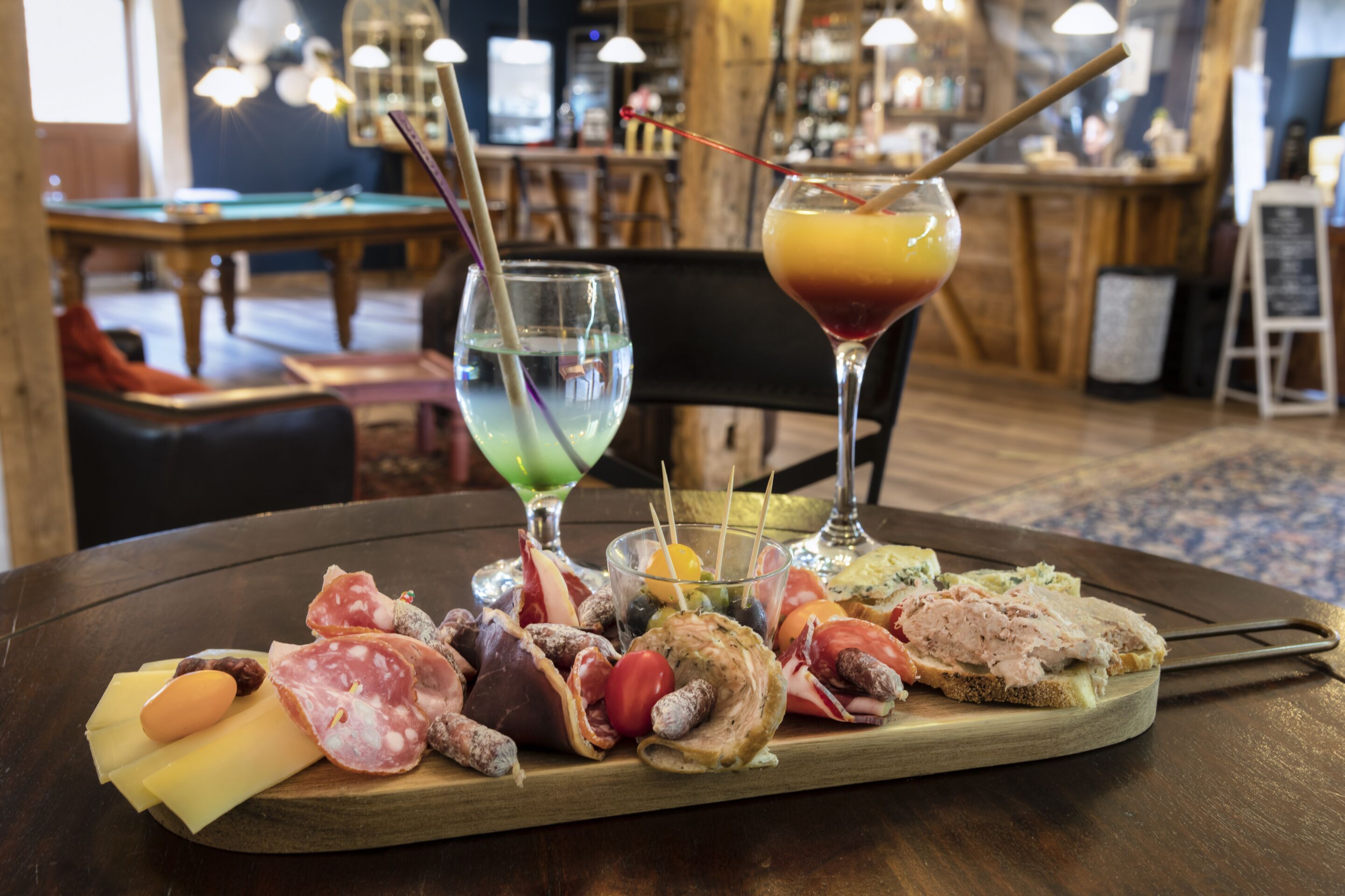 Una mesa de madera con una tabla de embutidos, dos bebidas y una zona de bar al fondo.