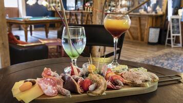 Una mesa de madera con una tabla de embutidos, dos bebidas y una zona de bar al fondo.