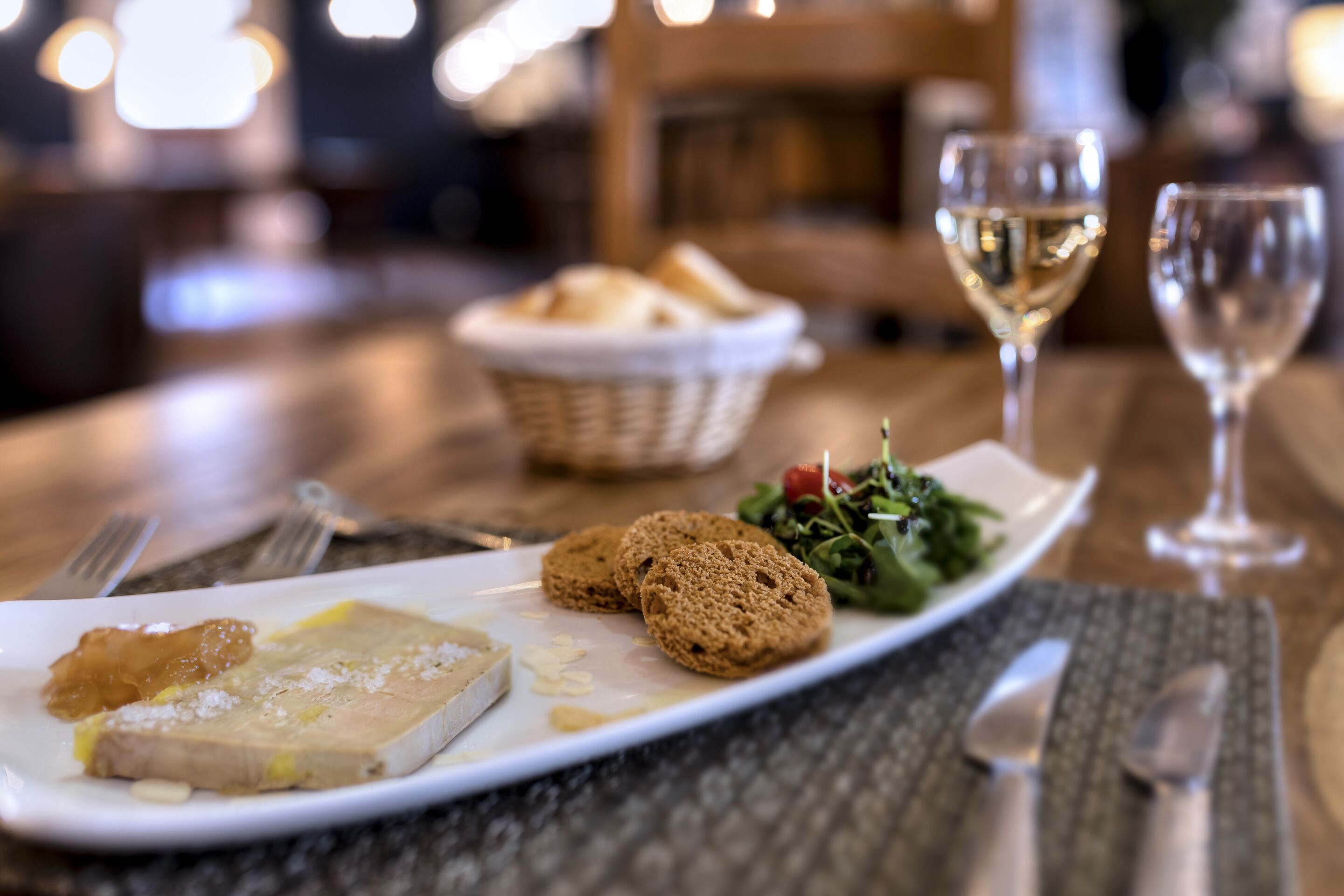 Un tavolo da pranzo con un piatto di cibo, due bicchieri di vino bianco e un cestino di pane.