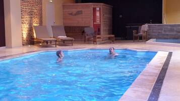 A swimming pool with two people in it, surrounded by lounge chairs and a brick wall.