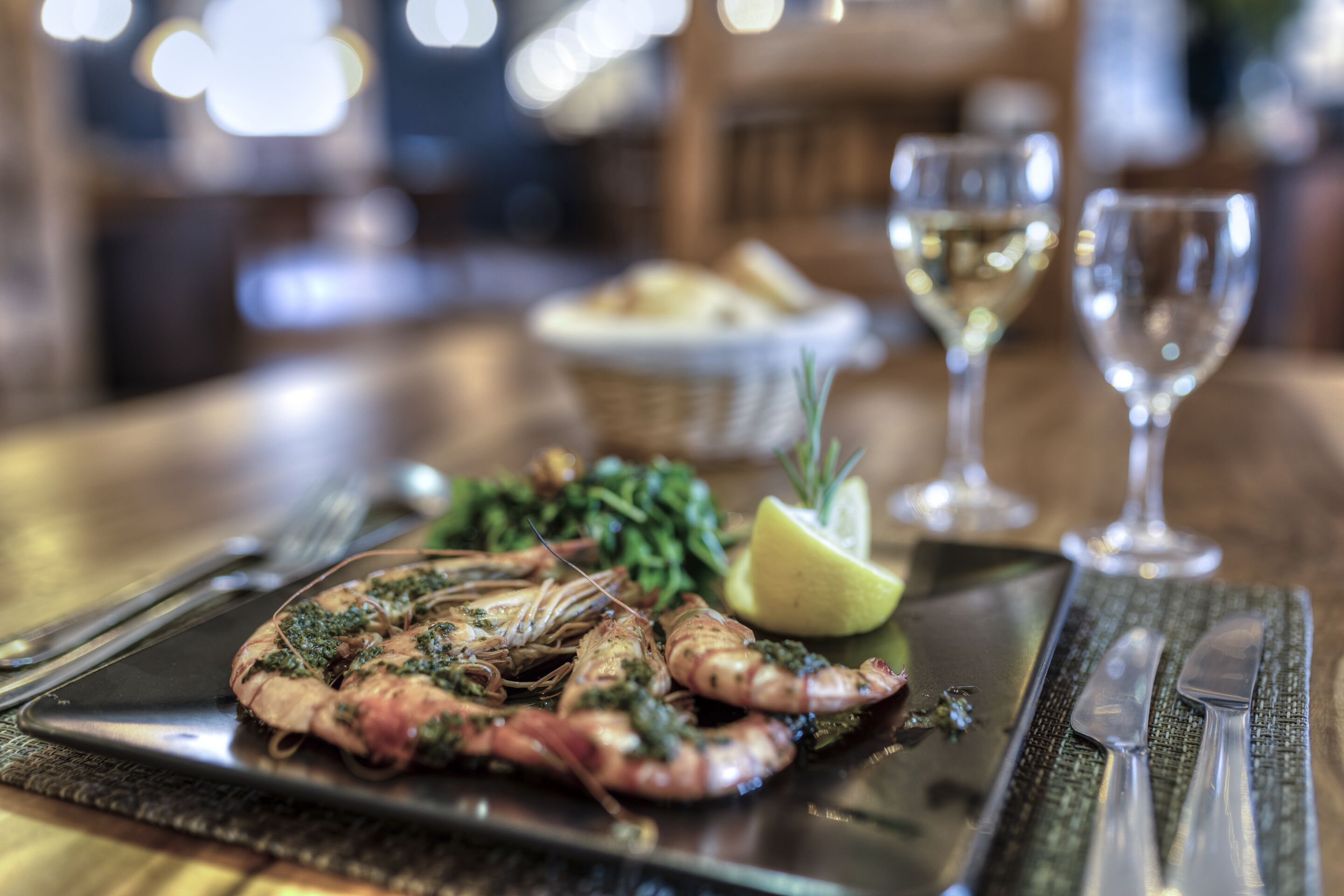Un plat de fruits de mer présenté dans une assiette, accompagné d’une tranche de citron et de quelques légumes verts, ainsi qu’d’un verre de vin blanc et d’une portion de pain sur la table.
