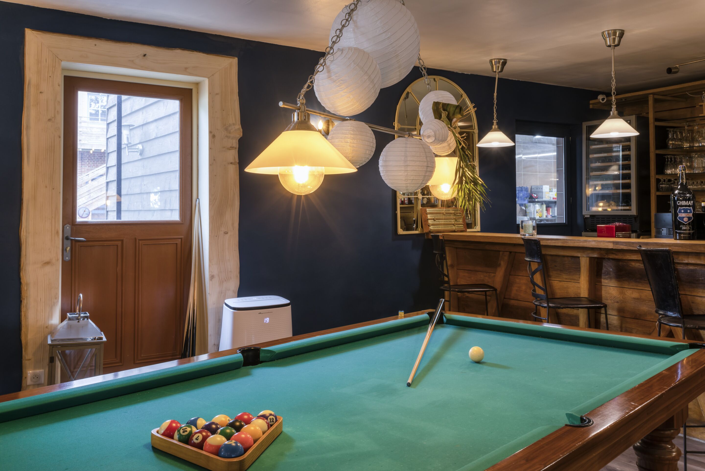 Uma mesa de bilhar com bolas, um bar com banquetas e uma janela com vista para edifícios.