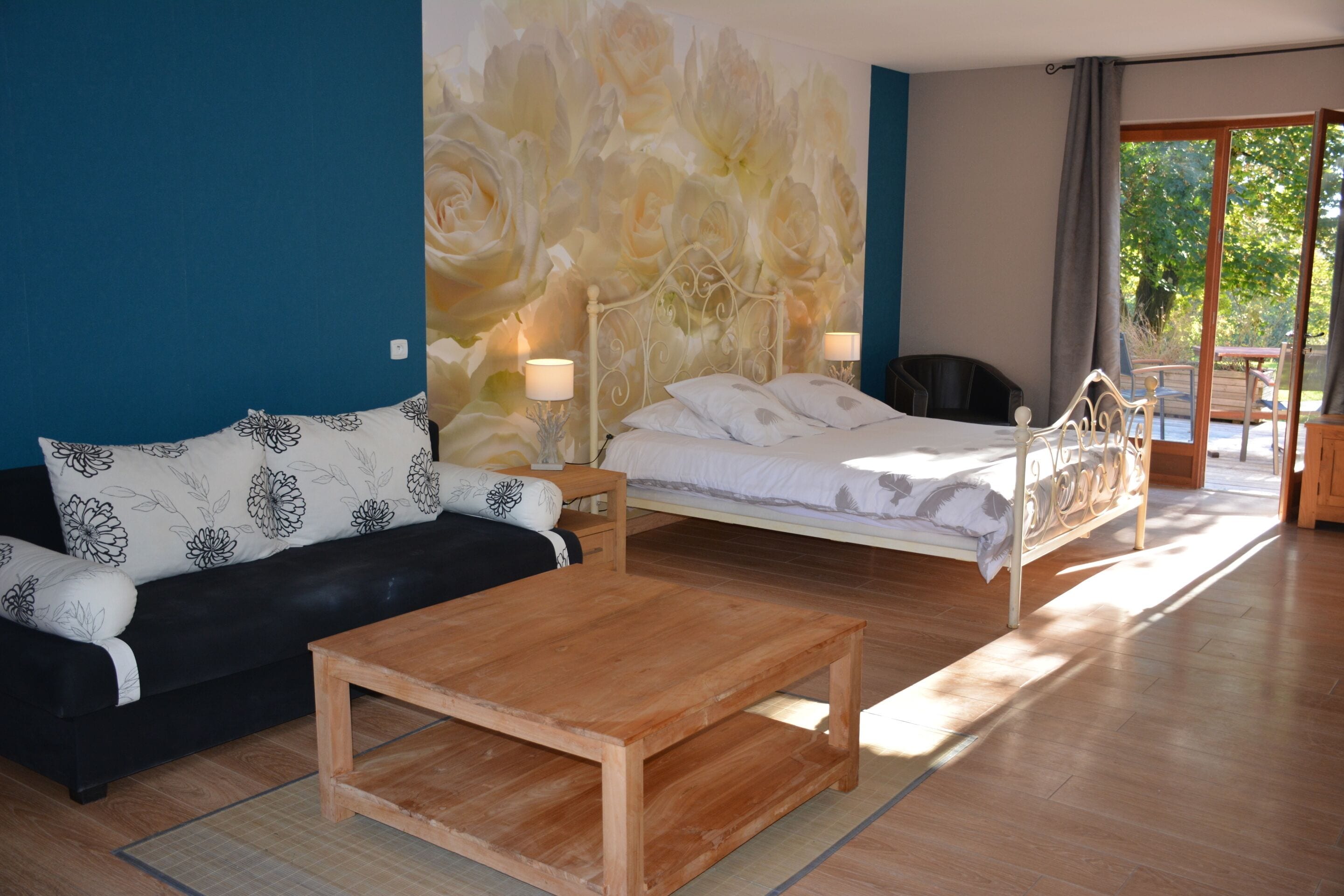 A bedroom with a bed, a wooden coffee table, a black sofa with floral cushions, and a large floral mural on the wall.