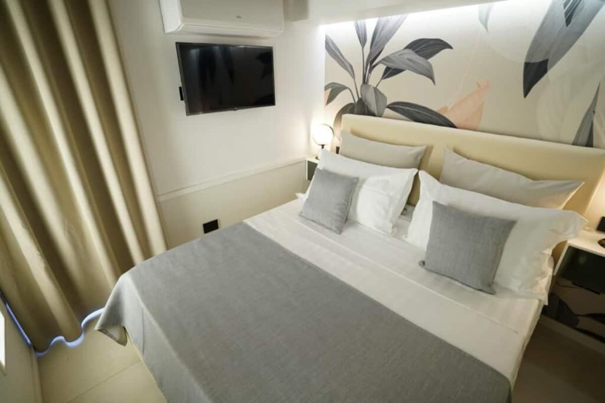 A neatly made bed with a grey and white color scheme, a flat-screen TV mounted on the wall, and a large plant mural on the wall behind the bed.