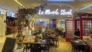 L’intérieur d’un restaurant, avec ses tables et ses chaises, un panneau néon indiquant « Le Monde Steak », et une composition florale décorative.