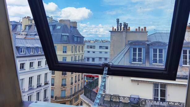 Vista desde una ventana que da a un paisaje urbano con varios edificios, incluyendo uno con balcón y otro con un letrero que dice "Optique".