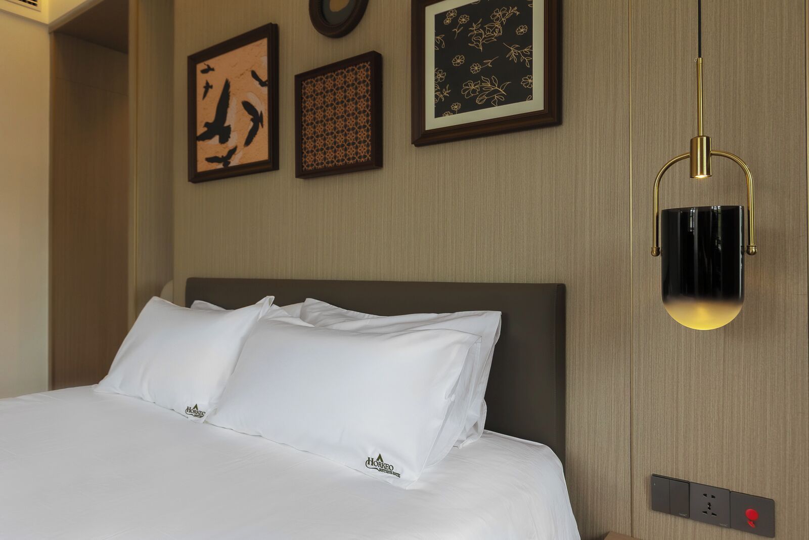 A hotel room with a neatly made bed, framed artwork on the wall, and a modern light fixture.