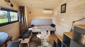 Interior acogedor de una cabaña con un sofá, una mesita con una botella, un banco y una ventana con cortinas.