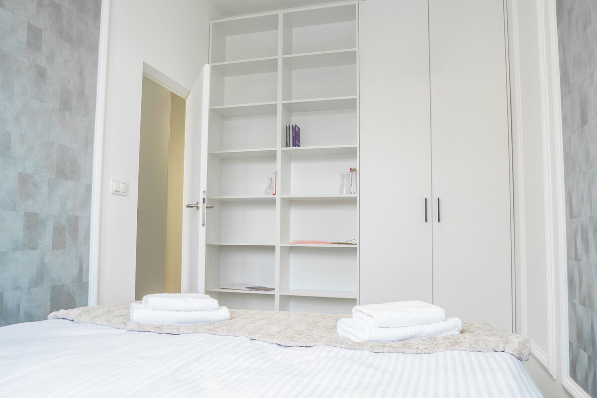 Un dormitorio con una cama, un armario blanco y una estantería con libros y objetos decorativos.
