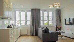 A modern kitchen with a grey sofa and a small table, large windows with curtains, and a flat-screen TV on the wall. - Długa 17 Residence (Kraków)
