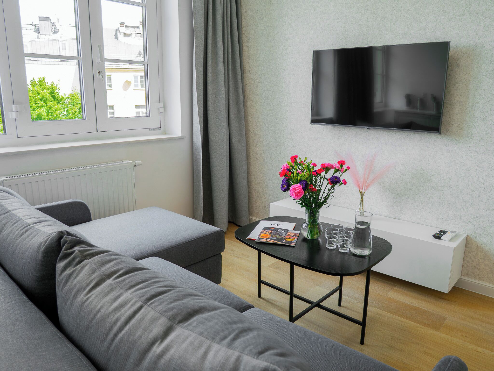 A modern living room with a grey sofa, a black coffee table with a magazine, a vase with flowers, a TV on the wall, and a window with curtains.