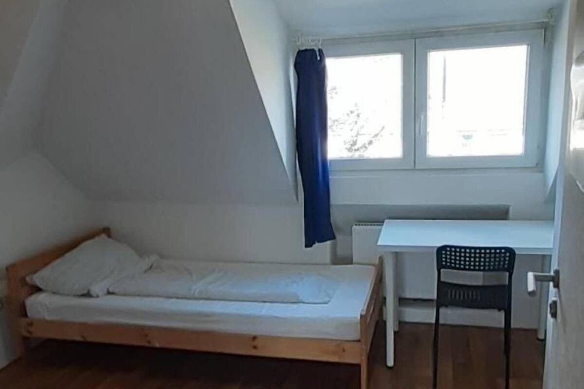 Un petit appartement d’une seule pièce, avec un plafond en pente, un plancher en bois, un lit, un bureau et une fenêtre avec des rideaux.