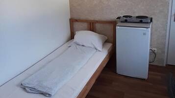 Um quarto pequeno com uma cama de solteiro, uma kitchenette modesta e um armário branco sobre o balcão.