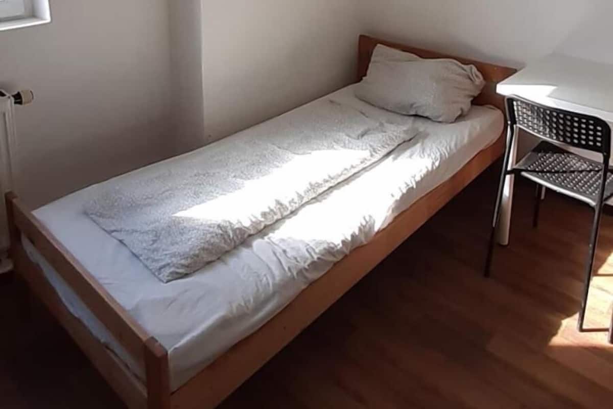 Une petite chambre avec un lit simple, un plancher en bois, un bureau blanc et une chaise.