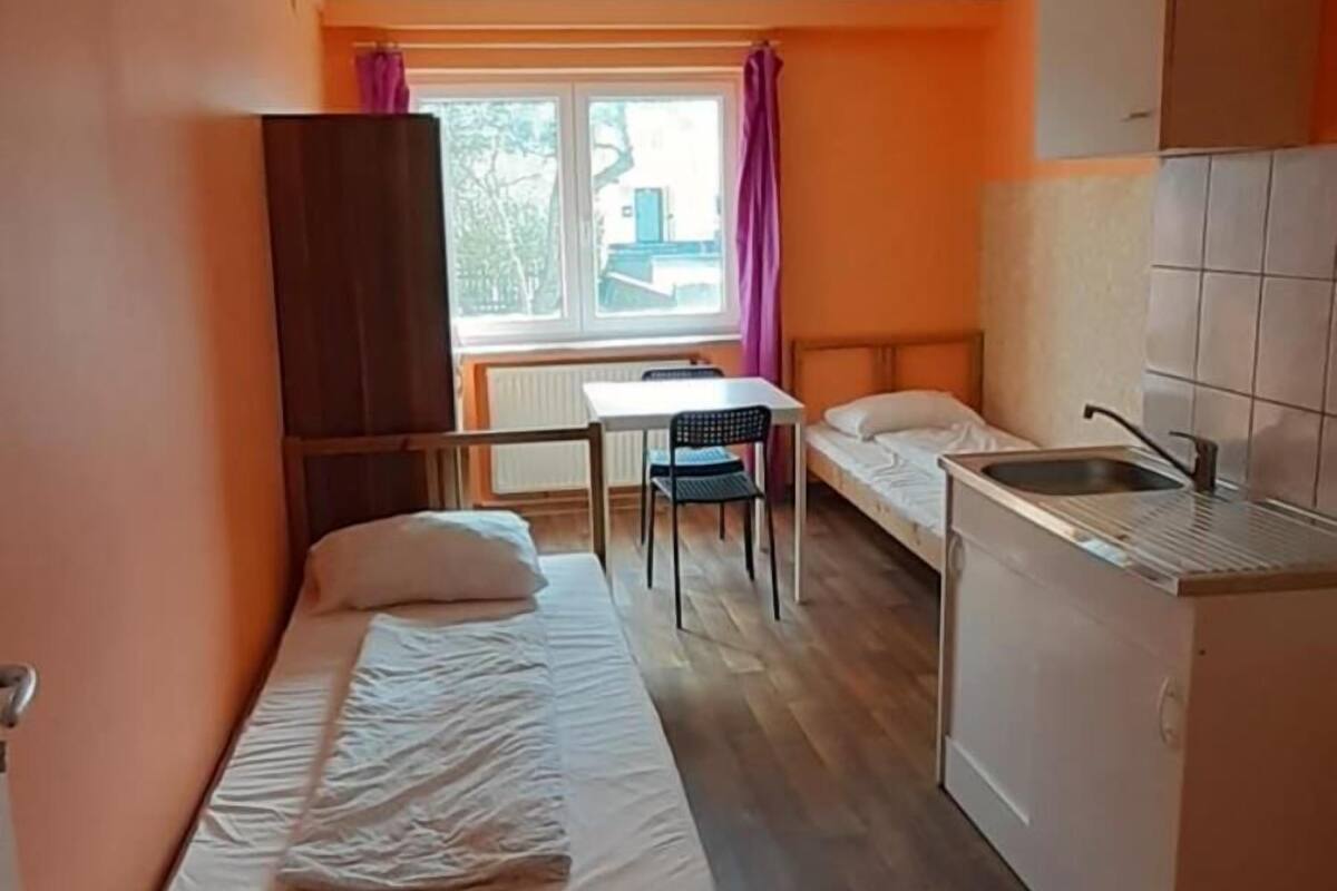 Une pièce de taille modeste comprenant un lit, une petite kitchenette, une fenêtre avec des rideaux et une vue sur l’extérieur.