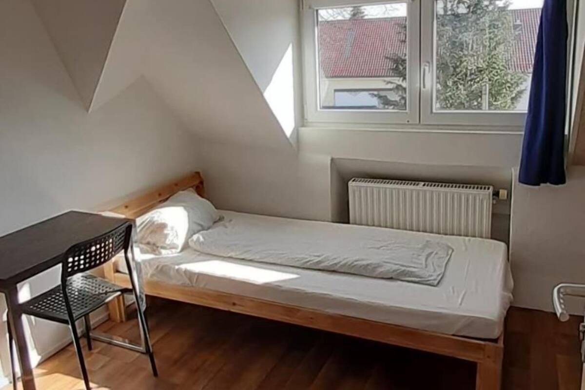 Une petite pièce bien éclairée, avec un plafond en pente, un lit simple, une petite table et une fenêtre donnant sur l’extérieur.