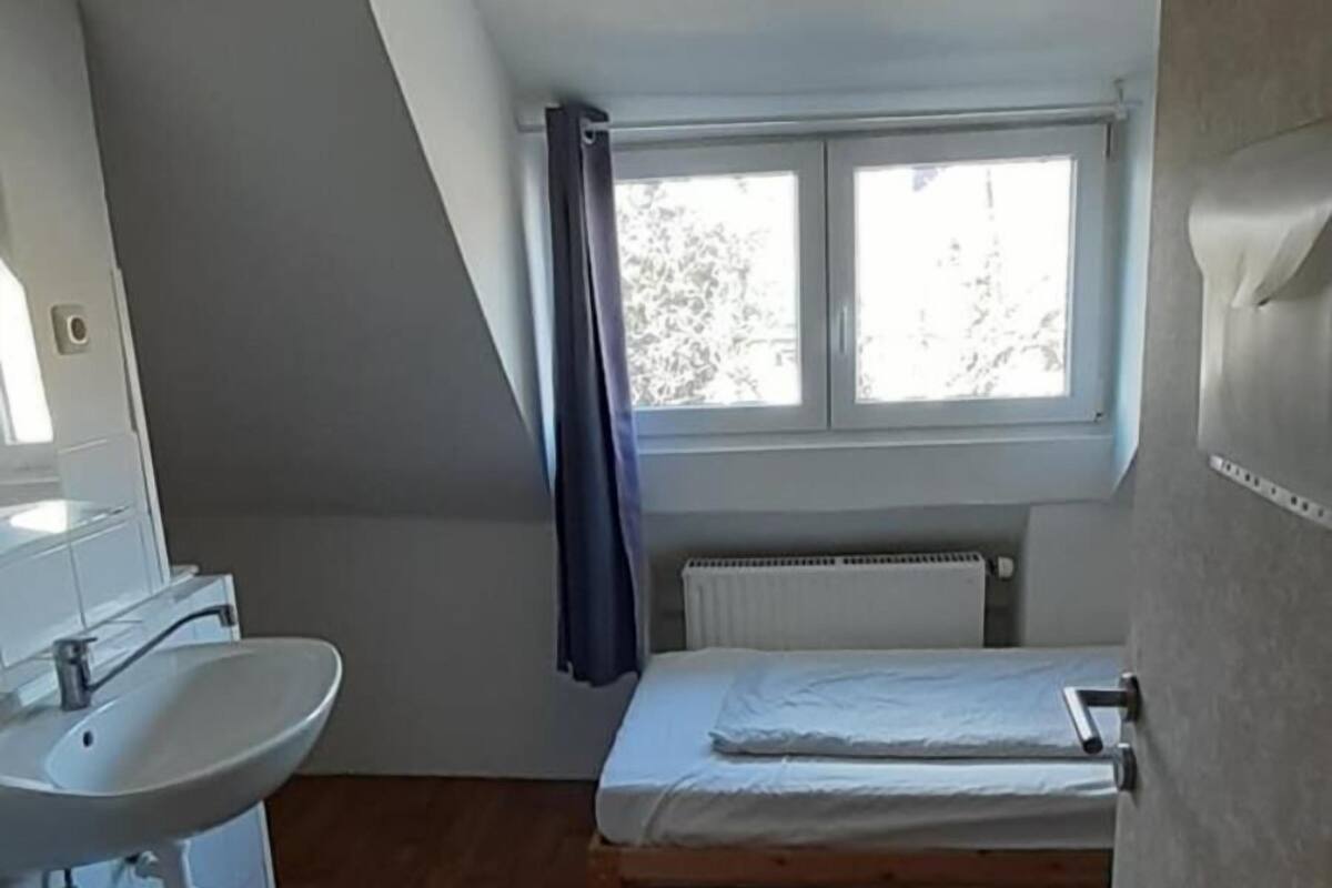 Une petite pièce étroite avec un plafond en pente, une fenêtre avec des rideaux, un lit, un lavabo et un radiateur.