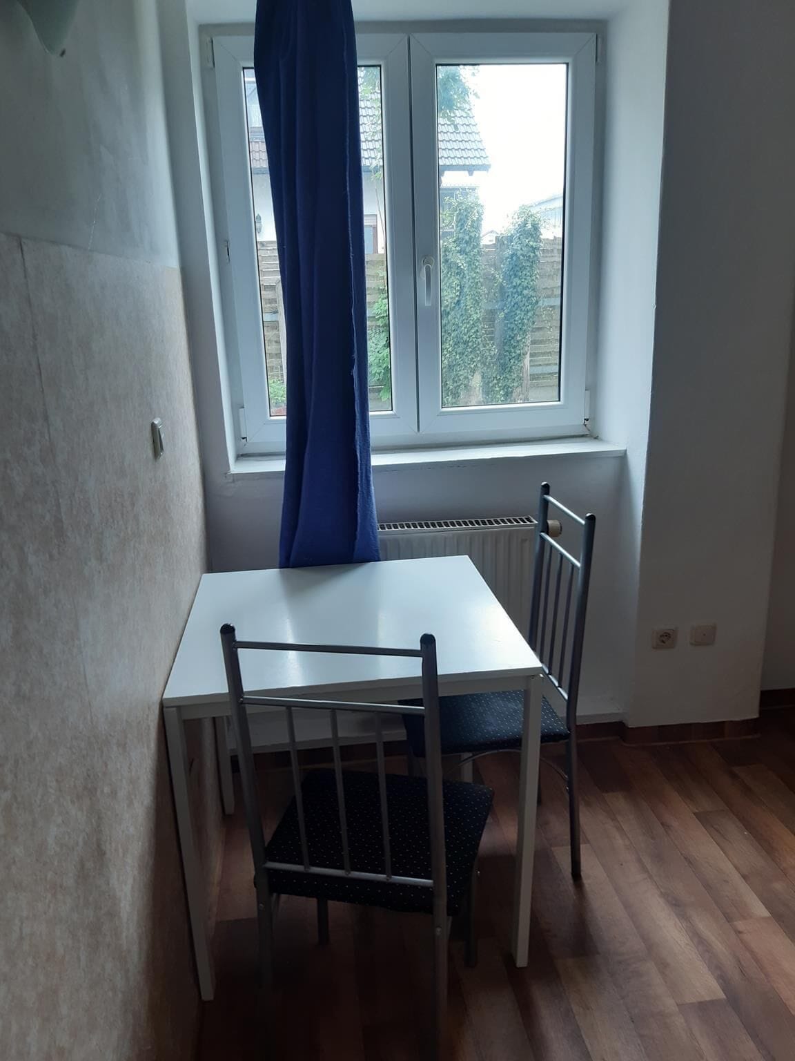 Ein kleines Zimmer mit einem weißen Tisch und zwei Stühlen, ein Fenster mit Blick auf Grünflächen und ein Heizkörper unter dem Fenster.