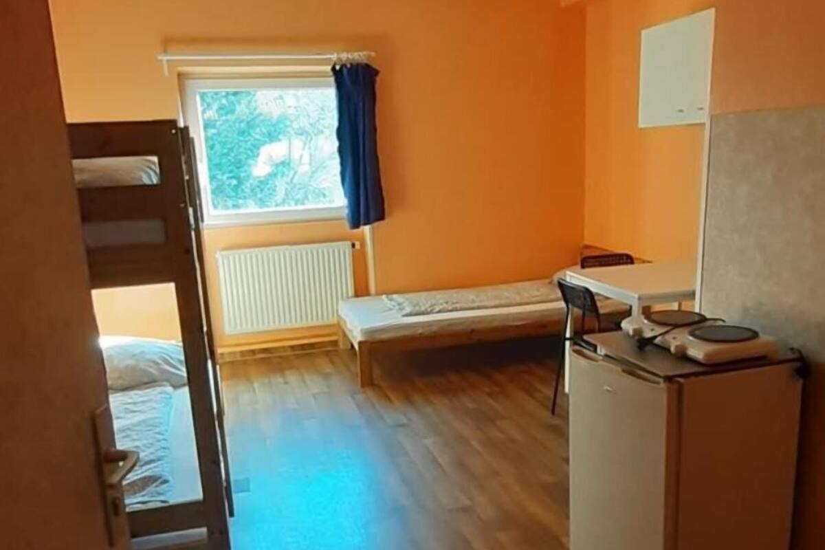 Une chambre avec un lit superposé, une petite table, un réfrigérateur et une fenêtre avec des rideaux.