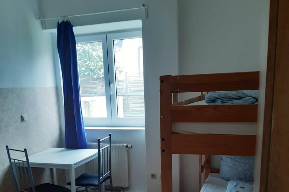 Une petite pièce bien éclairée, avec un lit superposé, un bureau avec une chaise et une fenêtre ornée de rideaux bleus.
