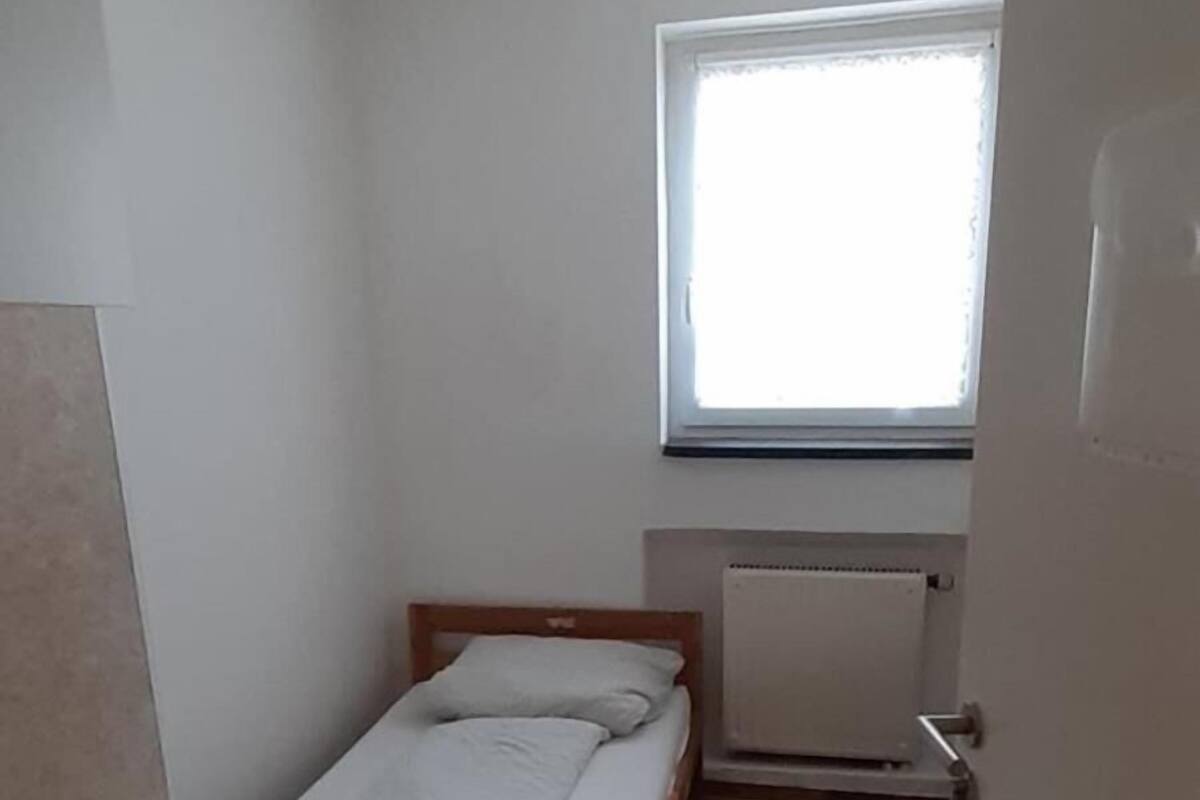 Une petite chambre avec un lit simple, un bureau et une fenêtre laissant entrer la lumière naturelle.