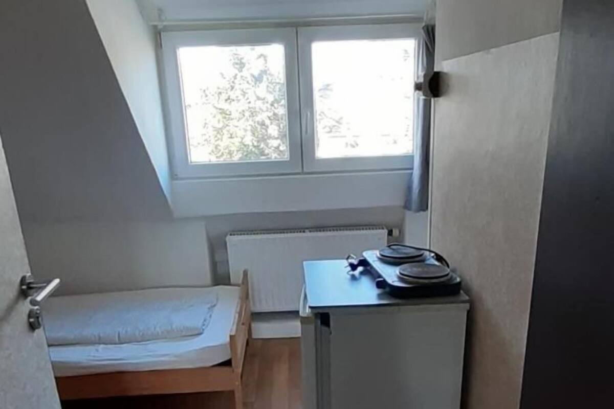 Une petite pièce étroite avec un plafond en pente, un lit, un radiateur et un bureau avec un ordinateur.