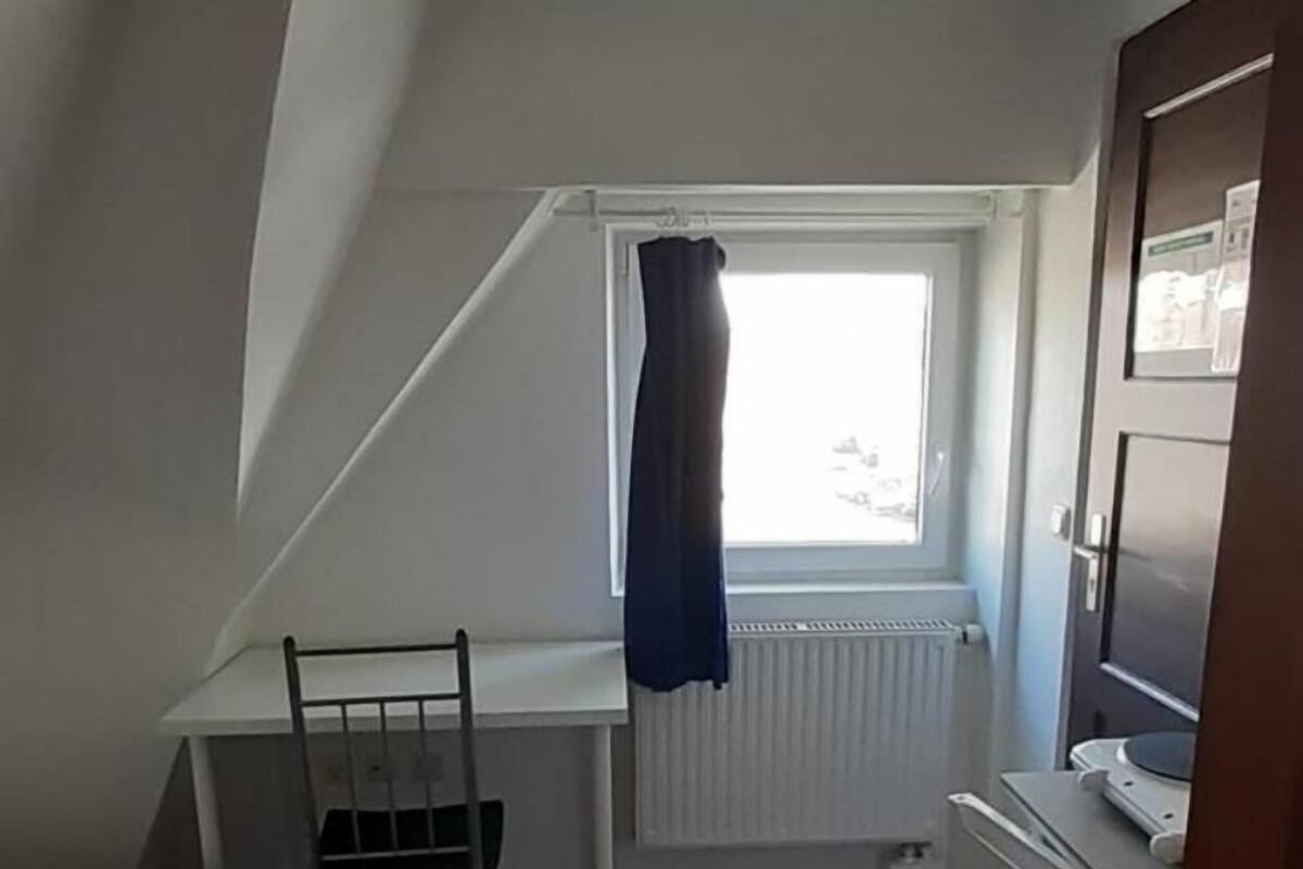 Une petite pièce aux dimensions réduites, avec un plafond en pente, un bureau blanc avec une chaise, un radiateur et un petit appareil électroménager blanc.