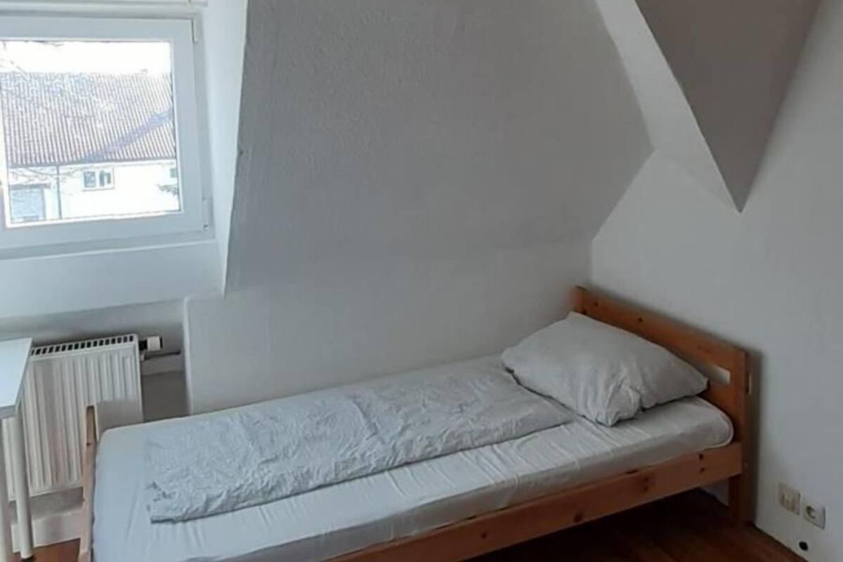 Une petite chambre avec un lit simple, un plafond en pente, un radiateur et une fenêtre.