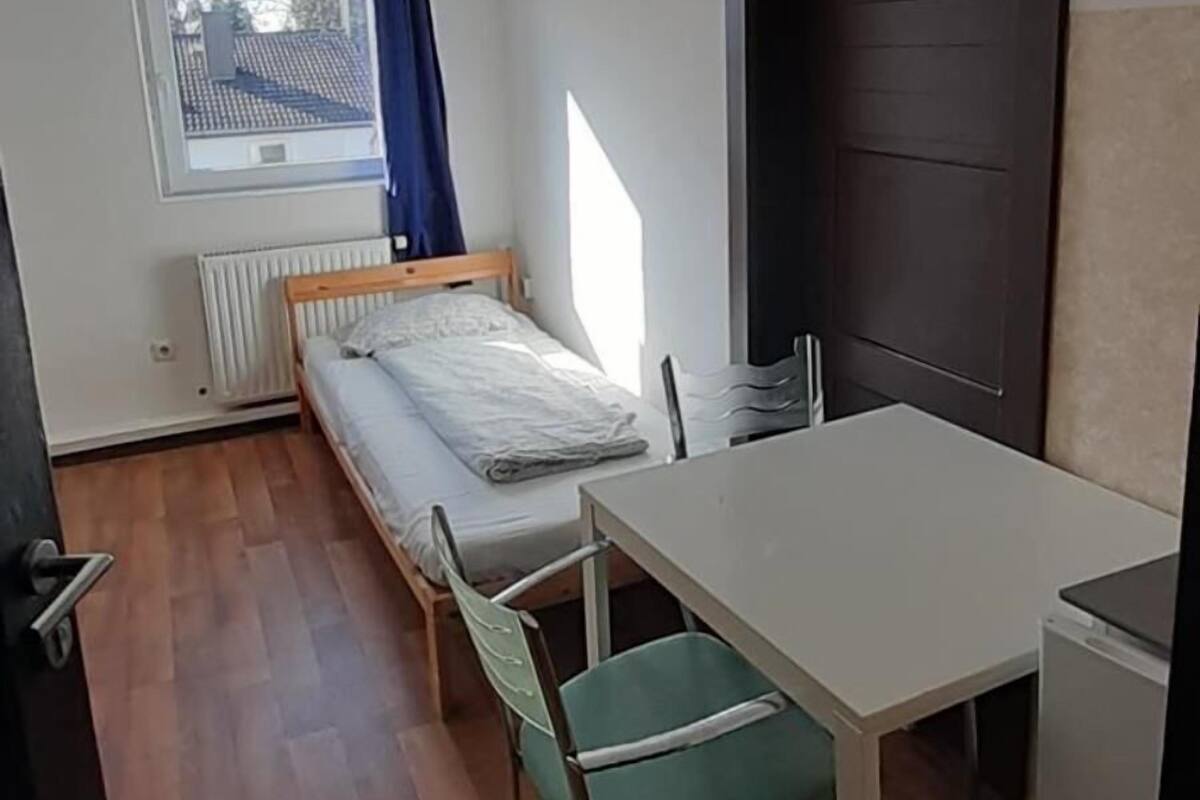 Une petite pièce, de taille modeste, comprenant un lit, une table et une chaise. Une fenêtre donne sur une maison, et un radiateur est placé en dessous.