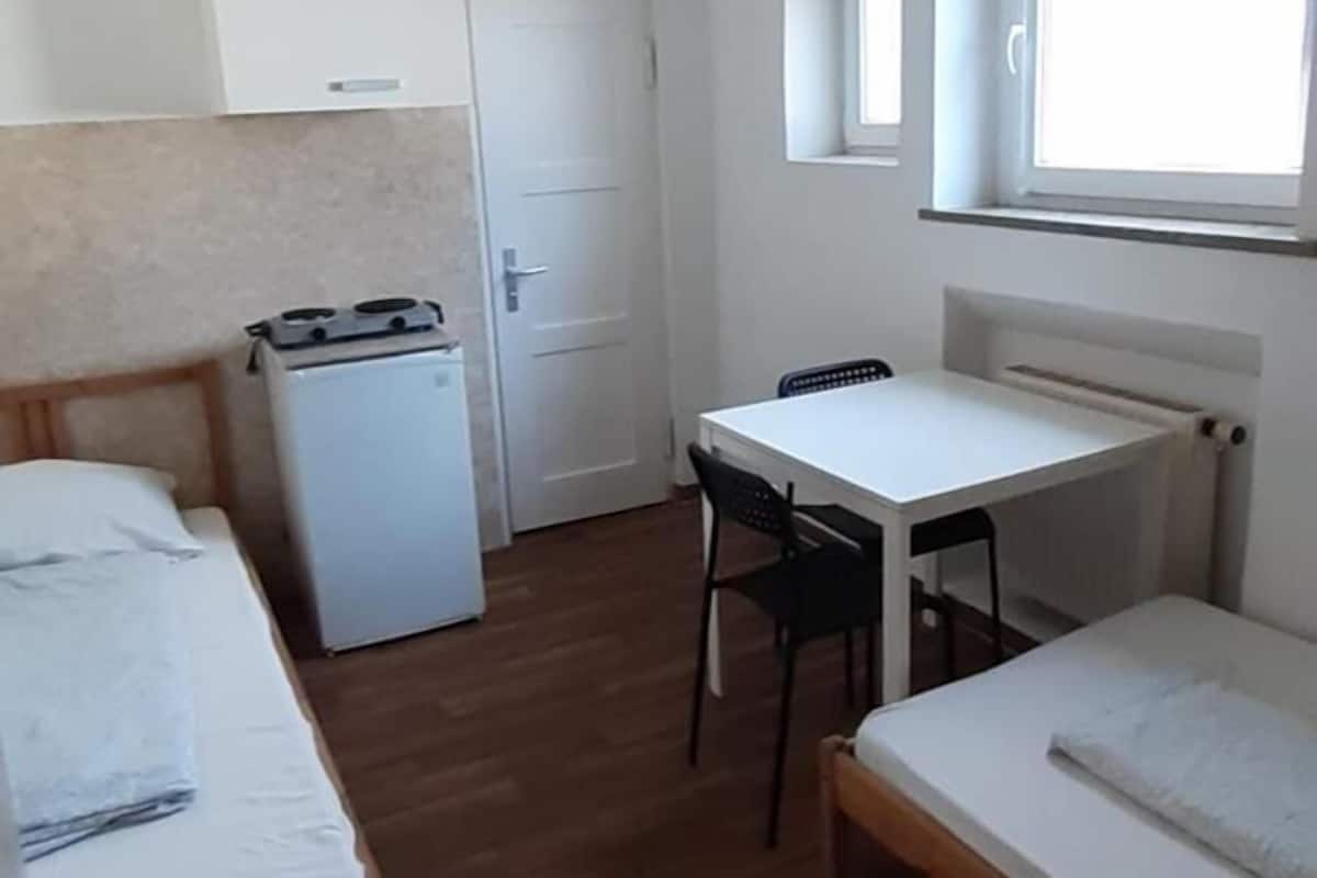 Une petite pièce compacte comprenant un coin cuisine, un lit simple, un bureau et une fenêtre.