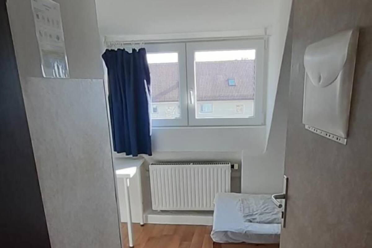 Un couloir étroit, doté d’un plancher en bois, mène à une pièce qui contient une fenêtre, un radiateur et un lit.