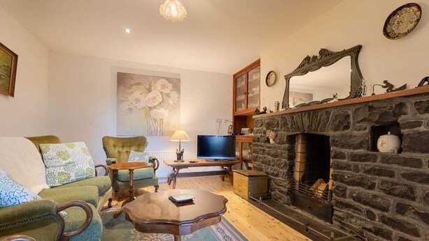 A cozy living room with a stone fireplace, green upholstered armchairs, a wooden coffee table, and a television. - Amazing Country Stay In County Galway, Ireland (Oughterard)