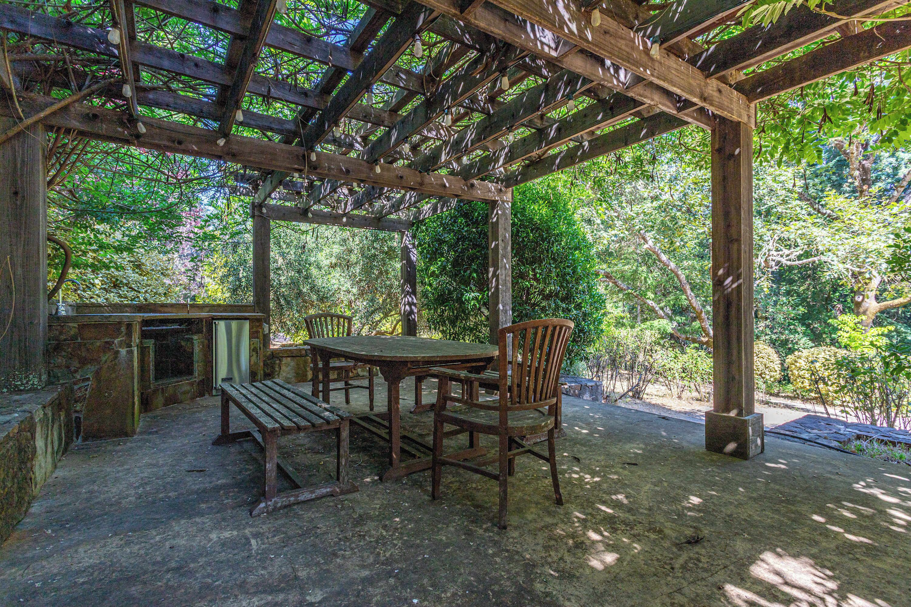 A wooden pergola structure with a table and chairs, surrounded by lush greenery.