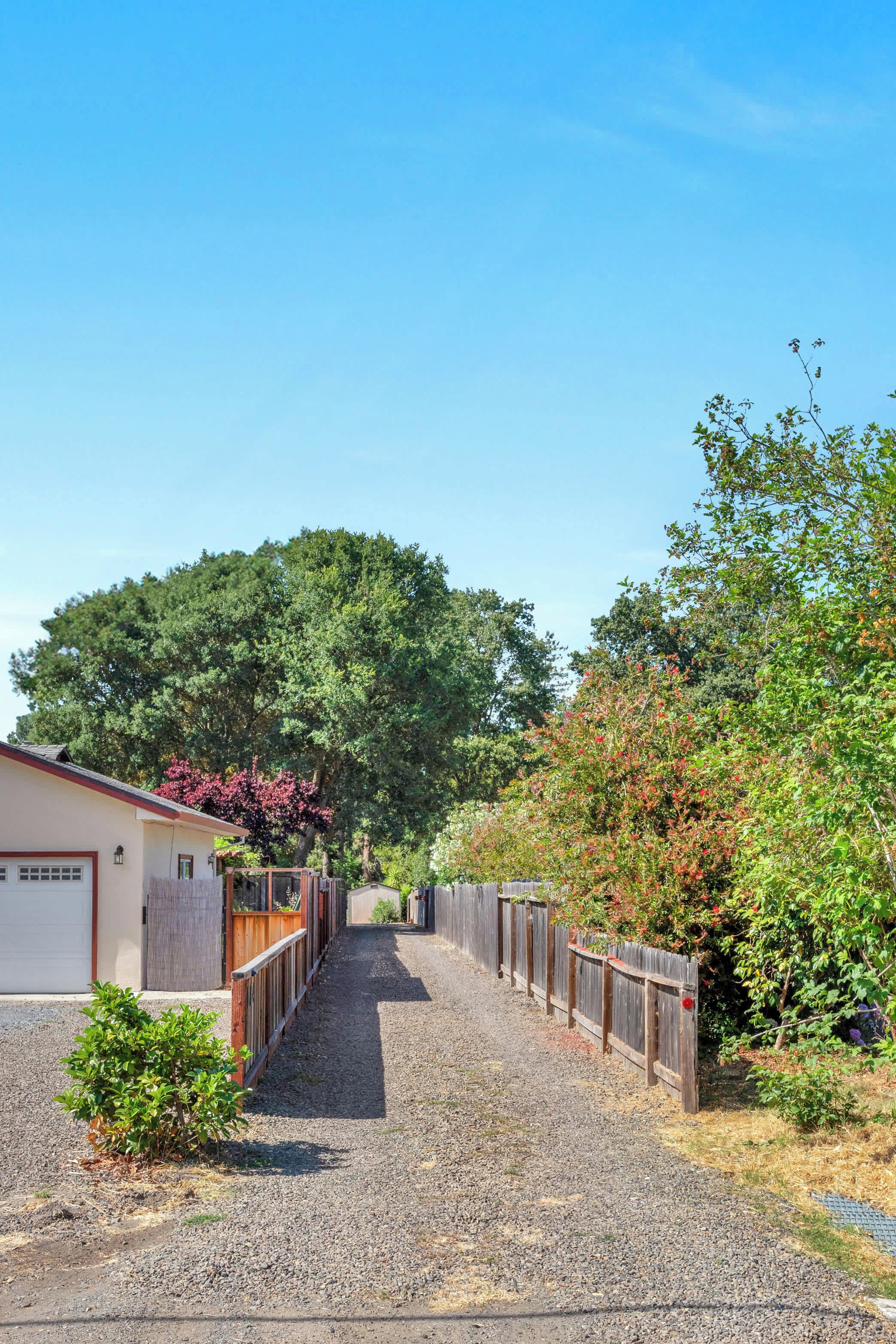 A gravel driveway leading to a house with a garage, surrounded by trees and a wooden fence.