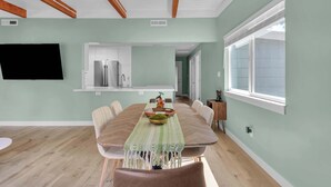 A dining area with a long table, chairs, a TV mounted on the wall, and a window with blinds. - Modern Country Home | Near Downtown & Bike Trail (Sebastopol)