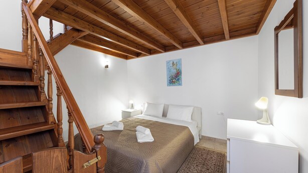 A bedroom with a wooden staircase, a bed with a brown bedspread, a white bedside table with a lamp, and a painting on the wall.
