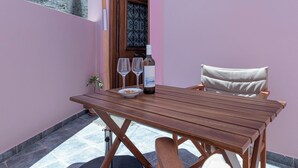 A wooden table with two wine glasses and a bottle, a cushioned chair, and a small potted plant on a balcony with a stone floor and a pink wall.