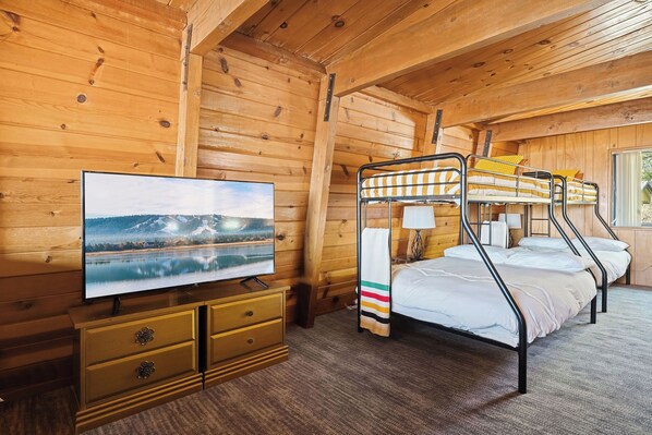 A wooden cabin room with bunk beds, a television on a wooden dresser, and a window with a view of a lake and mountains.