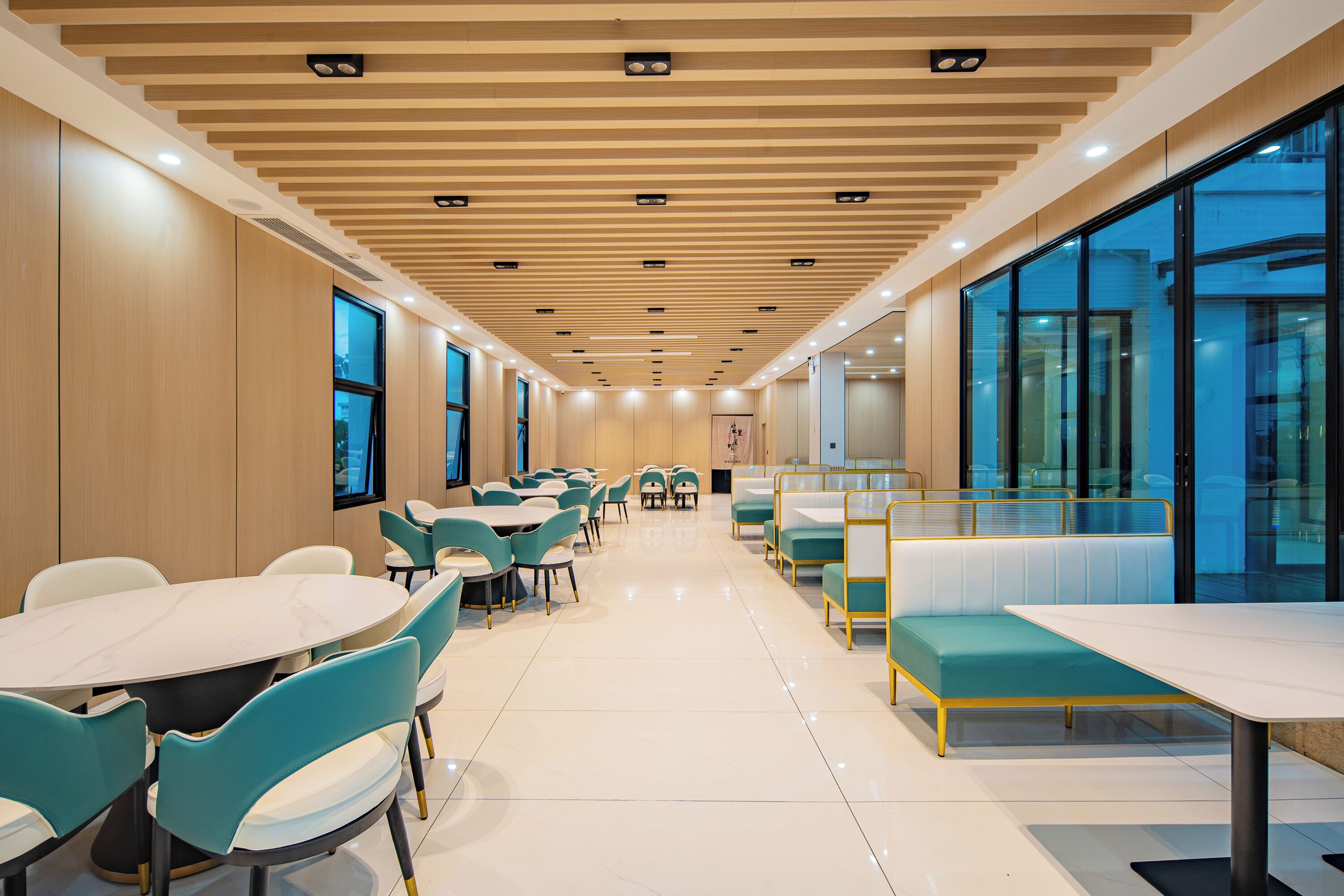 A modern dining area with round tables and teal chairs, wooden panel walls, and large windows.
