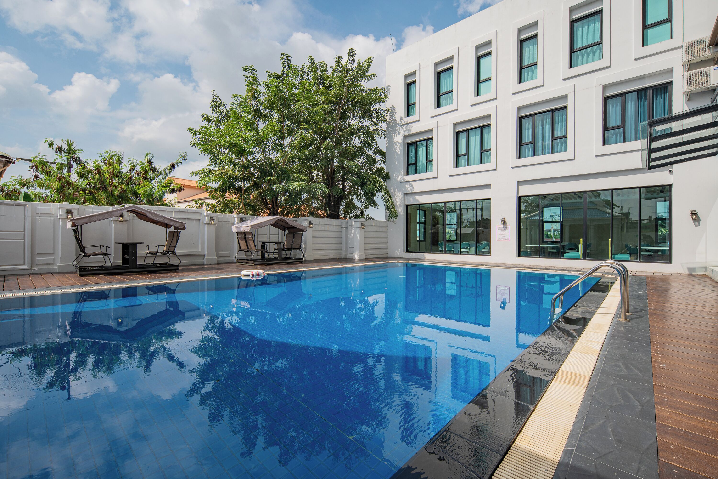 A swimming pool with a wooden deck, a white building with multiple windows, and a tree in the background.