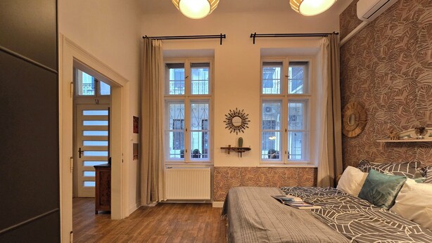 A bedroom with a bed, two windows, a radiator, a wall clock, and a ceiling light fixture.