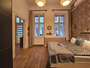 A bedroom with a bed, two windows, a radiator, a wall clock, and a ceiling light fixture. - Quiet Apartment in Kazinczy str. downtown (Budapest)