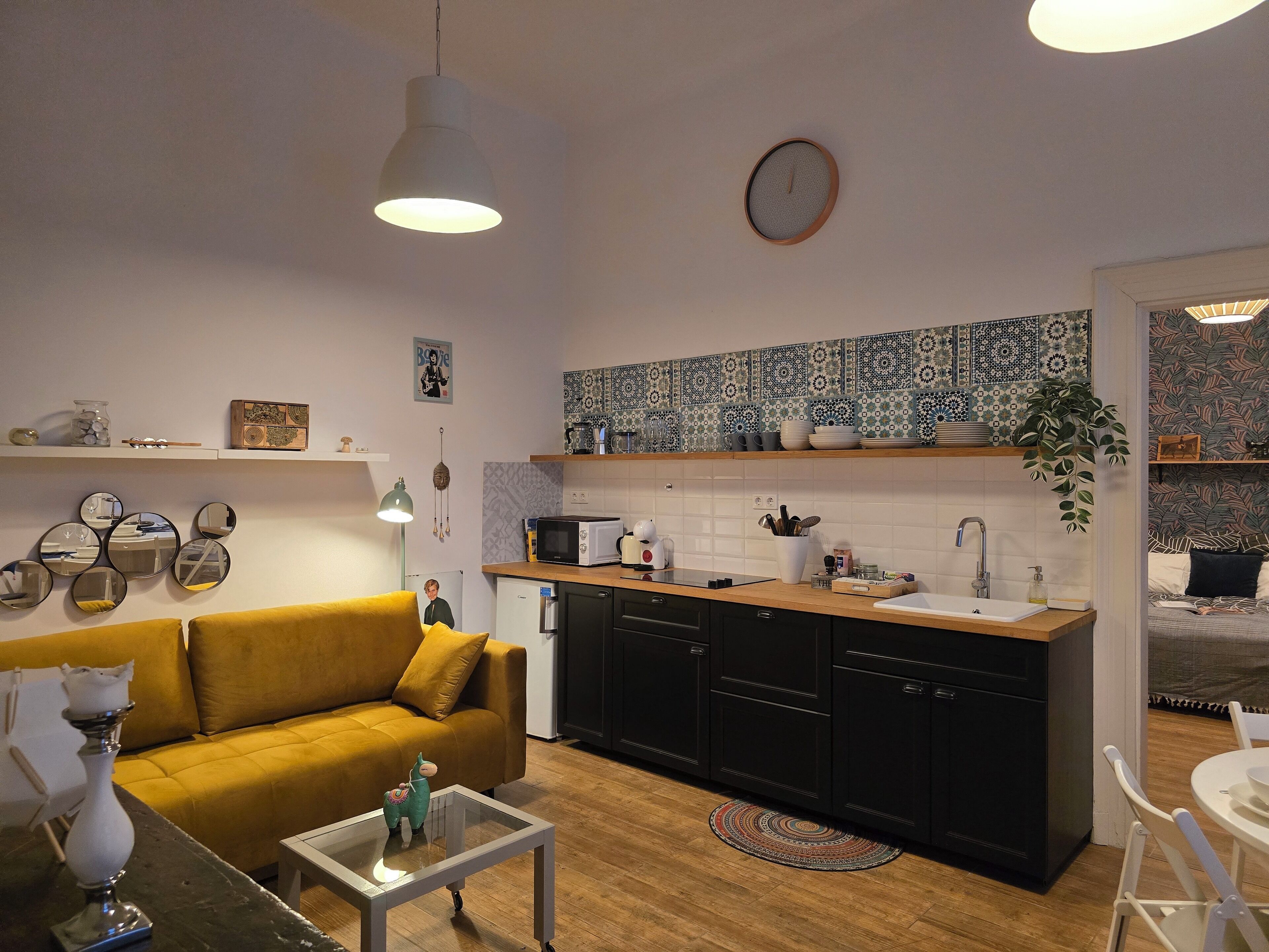 A modern kitchen with black cabinets, a wooden countertop, and a white sink. There is a yellow sofa and a glass coffee table in the living area.