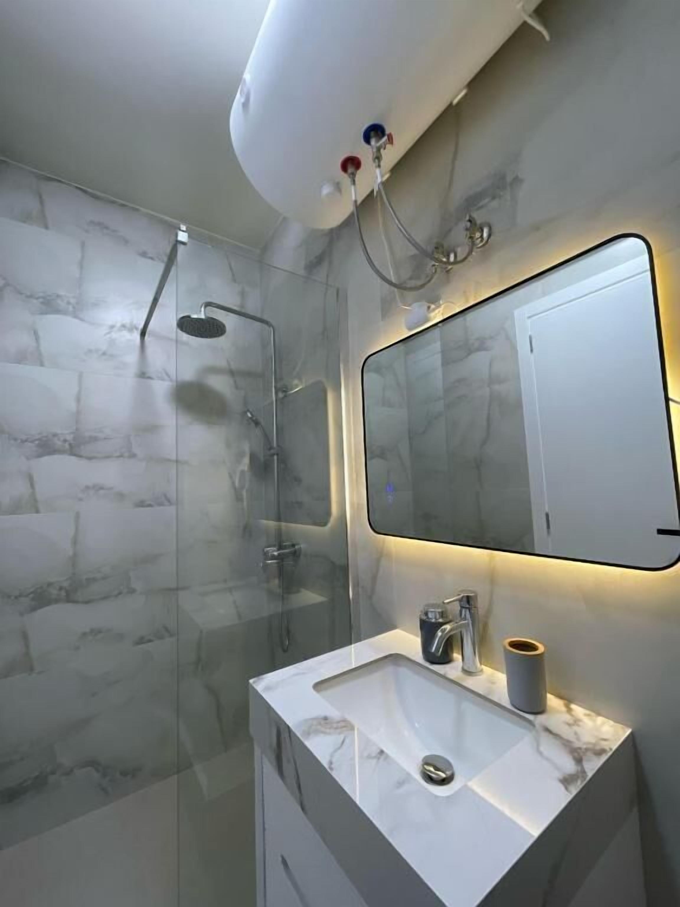 A modern bathroom with a glass-enclosed shower, a marble sink, and a mirror with integrated lighting.