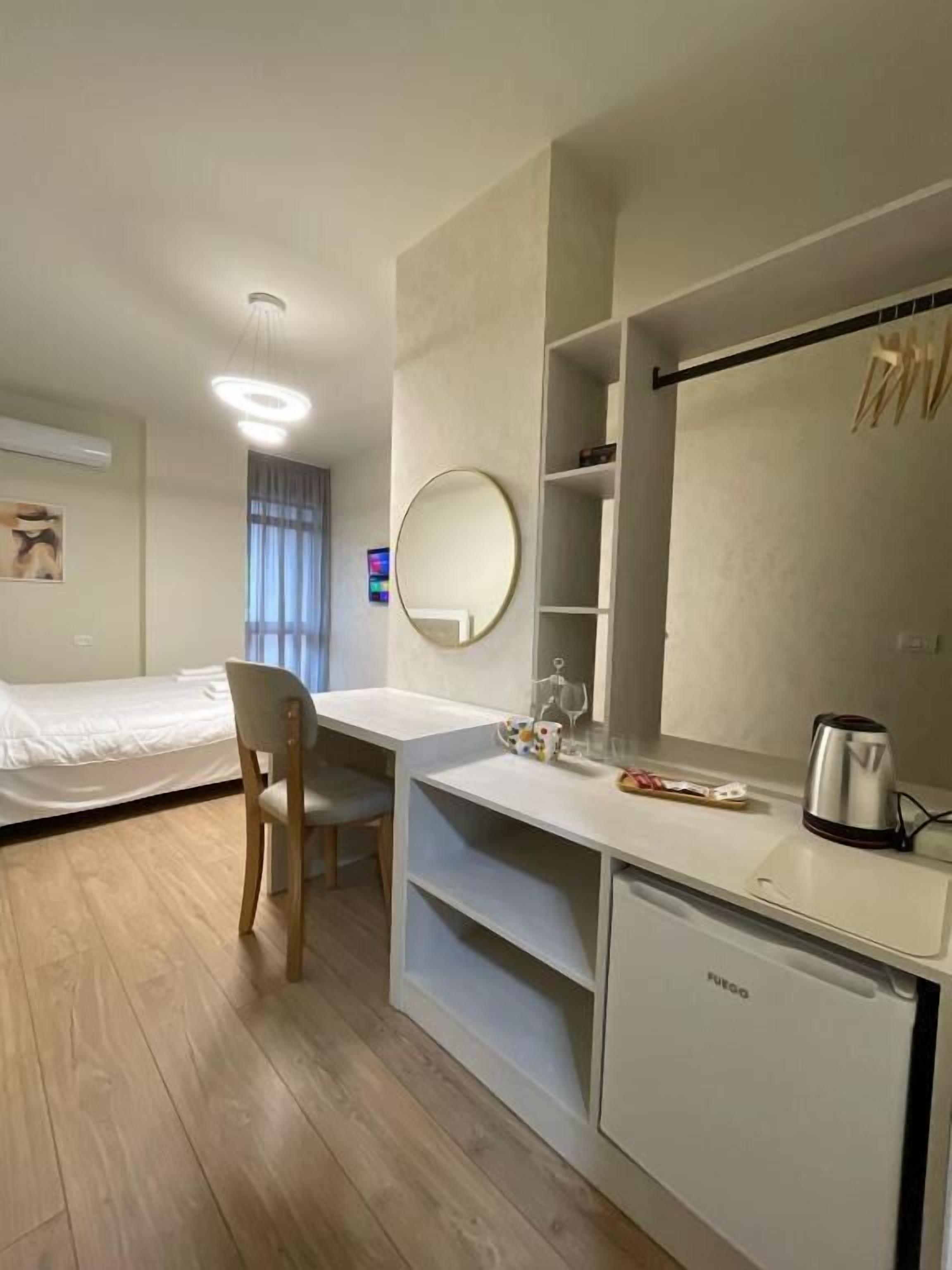 A modern hotel room with a wooden floor, a desk with a chair, a bed, a mirror, a shelf with a kettle, and a window with curtains.