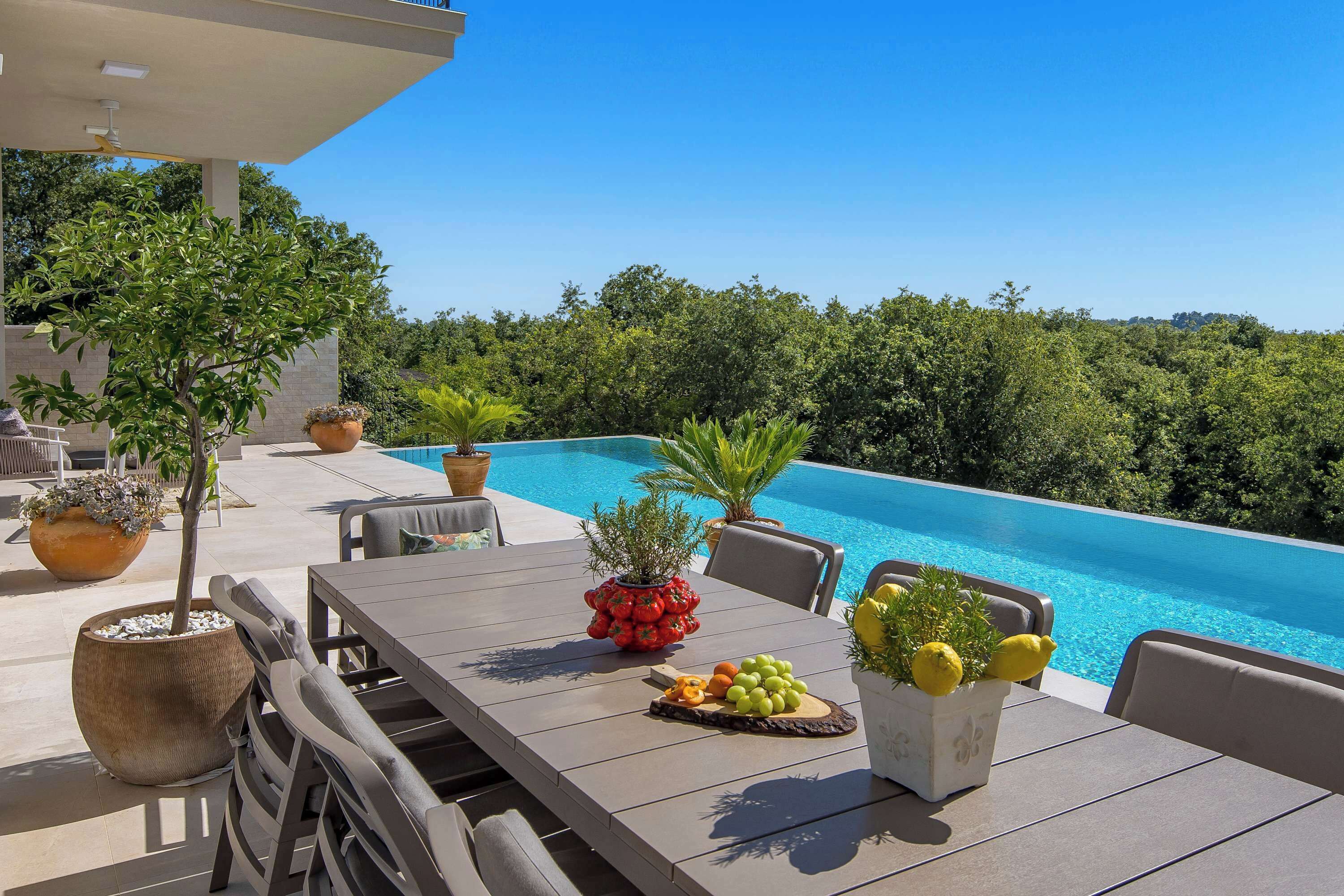 Un espace repas aménagé au bord de la piscine, avec une table dressée pour deux, entouré de plantes en pot et offrant une vue sur une piscine à l’eau d’un bleu limpide.