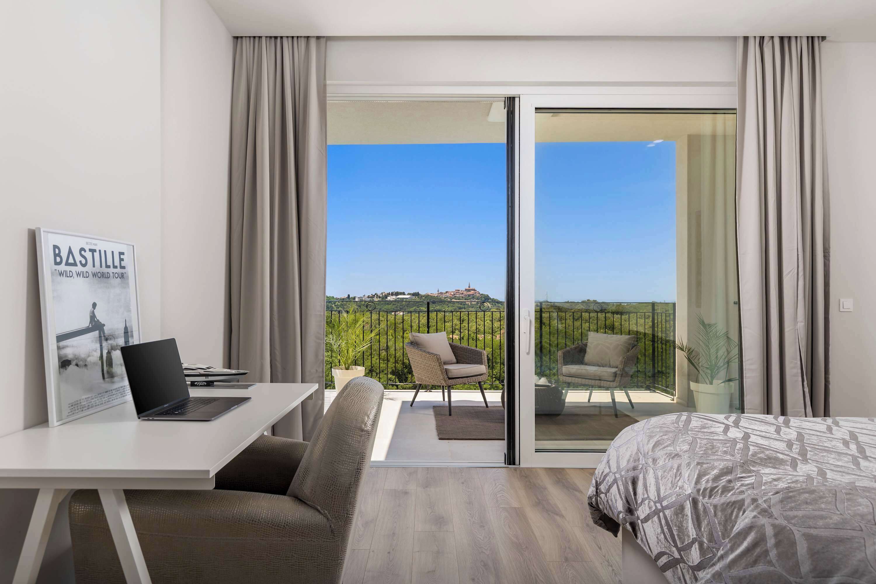 A modern hotel room with a bed, a desk with a laptop, a chair, and a balcony with a view of the countryside.