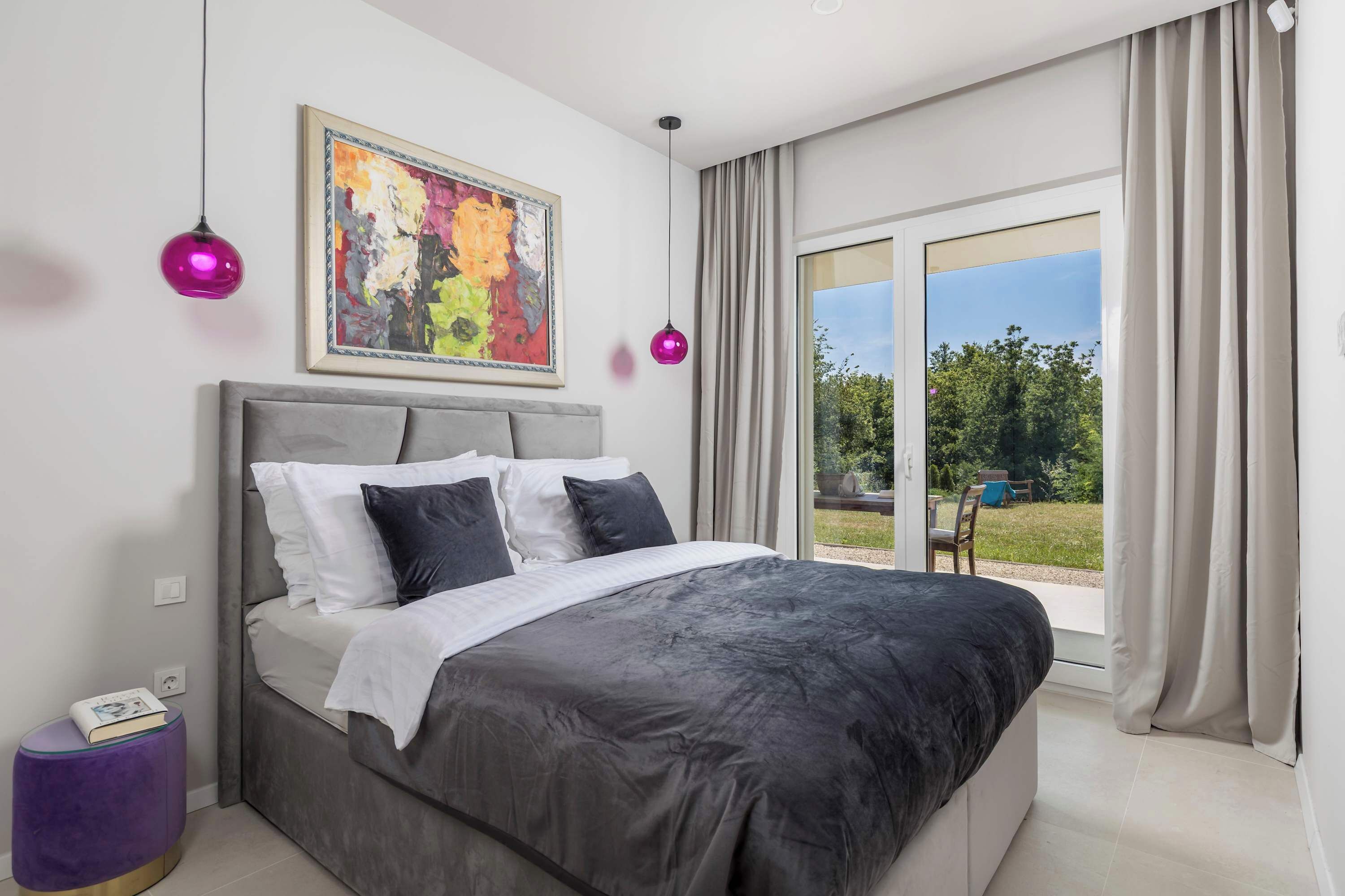 A bedroom with a bed, a window with curtains, a painting, and a purple lamp.