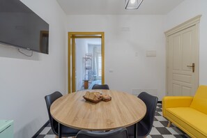 A room with a round wooden table, black chairs, a yellow sofa, a TV mounted on the wall, and a door leading to another room.